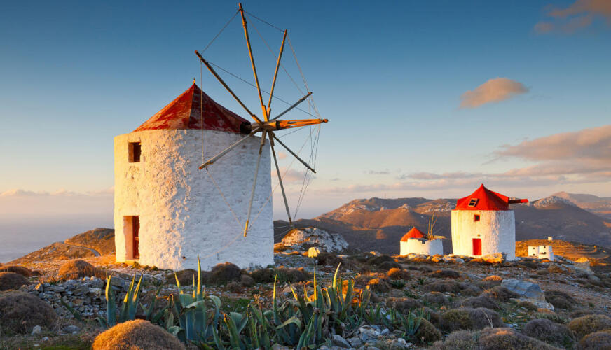 Λευκοί ανεμόμυλοι με κόκκινη στέγη στην κορυφή λόφων με αραιή θαμνώδη βλάστηση. Στο βάθος διακρίνονται και άλλοι λόφοι.