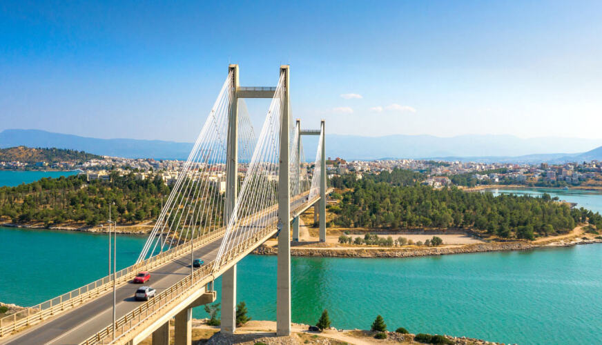 Μεγάλη σύγχρονη κρεμαστή γέφυρα πάνω από θάλασσα που οδηγεί σε δεντρόφυτη έκταση και στο βάθος μια πόλη.