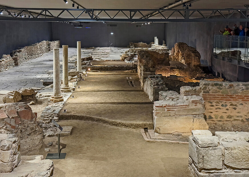 Θέα των αρχαίων ερειπίων που διατηρούνται σε εσωτερικό χώρο στο Μετρό της Θεσσαλονίκης, Σταθμός Βενιζέλου, με εμφανείς τοίχους, κίονες και ψηφιδωτά δάπεδα, ενώ επισκέπτες παρατηρούν από μια υπερυψωμένη διάβαση.




